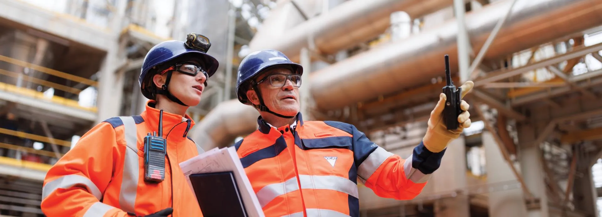 Two Vicat workers in hard hats and orange safety gear inside a cement plant